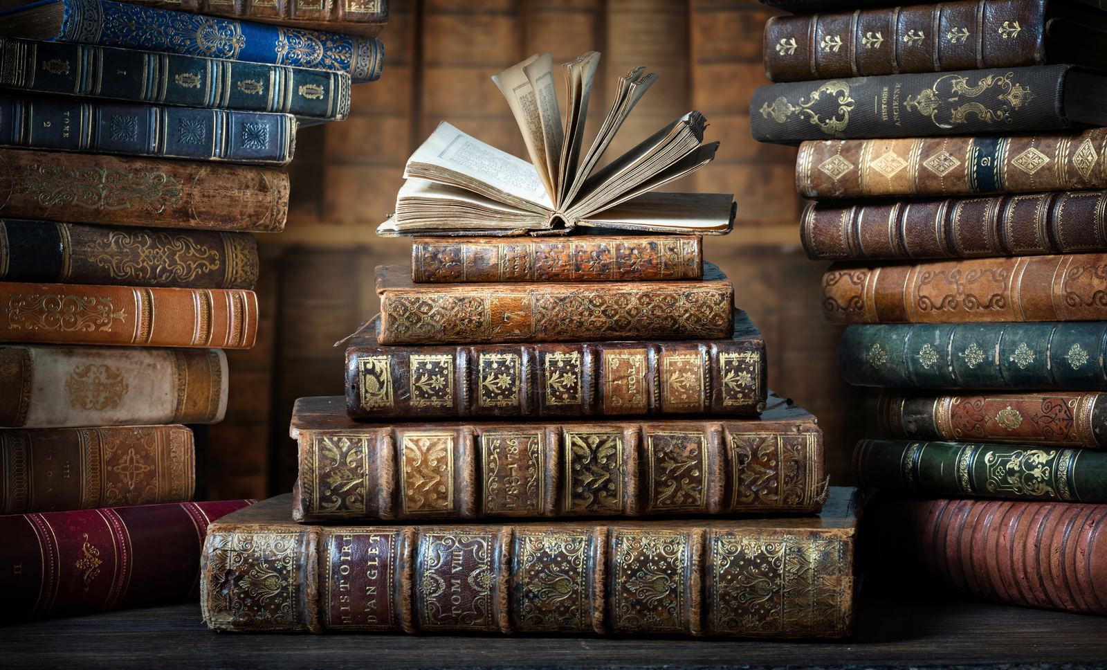 Stacks of antique leather-bound books with gilt spines, an old book lying open on top, in warm light